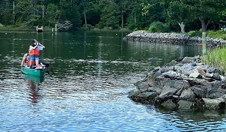 Two people in a canoe on calm water near a rock shoreline and trees.