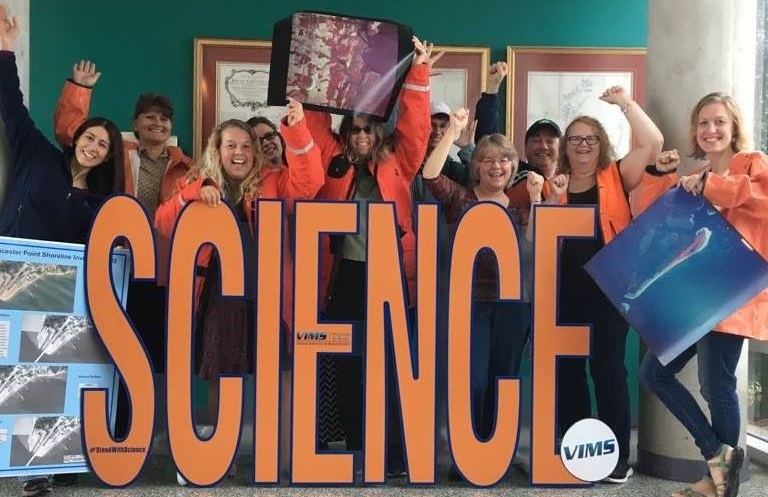 Group of people smiling and raising their arms behind large letters spelling “SCIENCE,” some holding posters and wearing orange jackets.