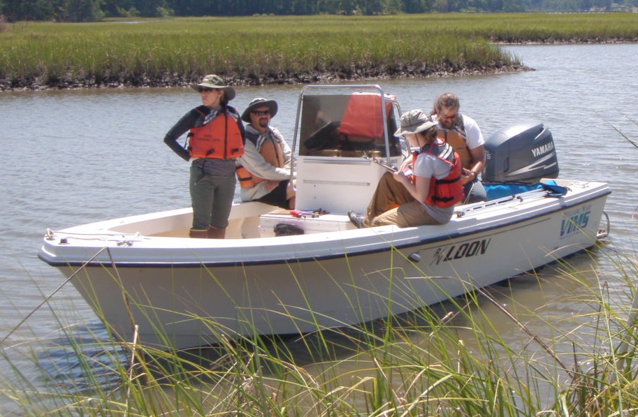 Group of people in life vests on a small boat in a marsh while another person stands on shore holding vegetation.