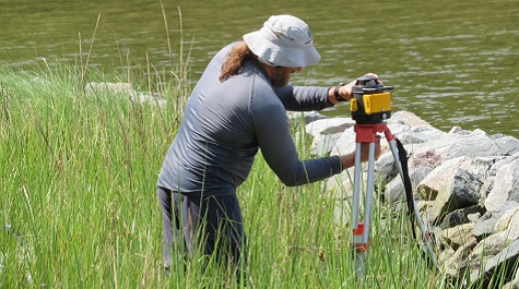 Person using data collection equipment on a tripod in tall grass along a shoreline.