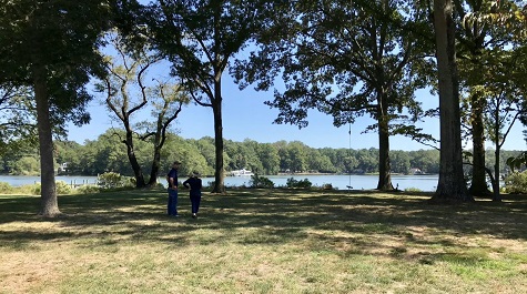 Two people walking across a grassy area with trees toward a waterfront in the distance.
