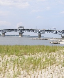 Grassy sand shoreline with a rock breakwater and a long bridge spanning across the water in the background.