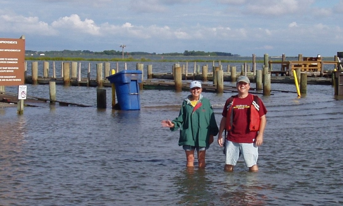 Two people standing in knee-deep floodwater at a marina with submerged docks and posts behind them.