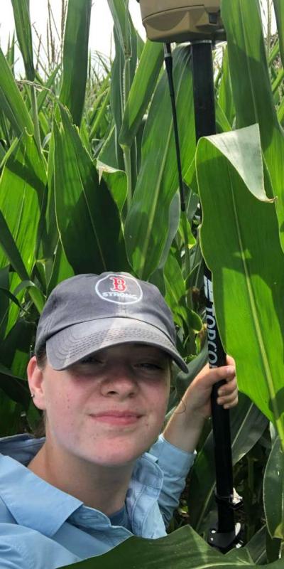 Grace Levins holds a GNSS survey pole among farmland crops.