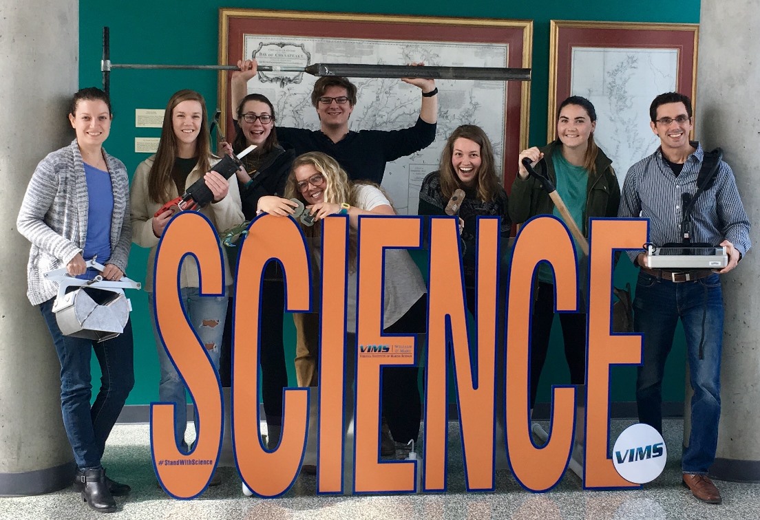 Coastal Geology lab group poses with field equipment around a large sign that says "SCIENCE"