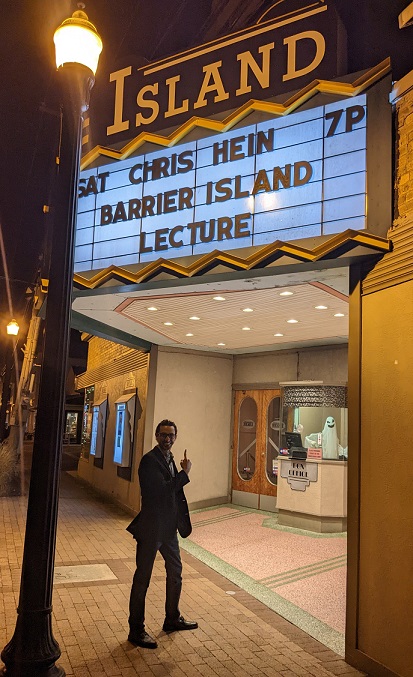 Chris Hein stands outside of Chincoteague Theater, pointing up at the marque, which reads "Chris Hein Barrier Island Lecture, Saturday 7pm"